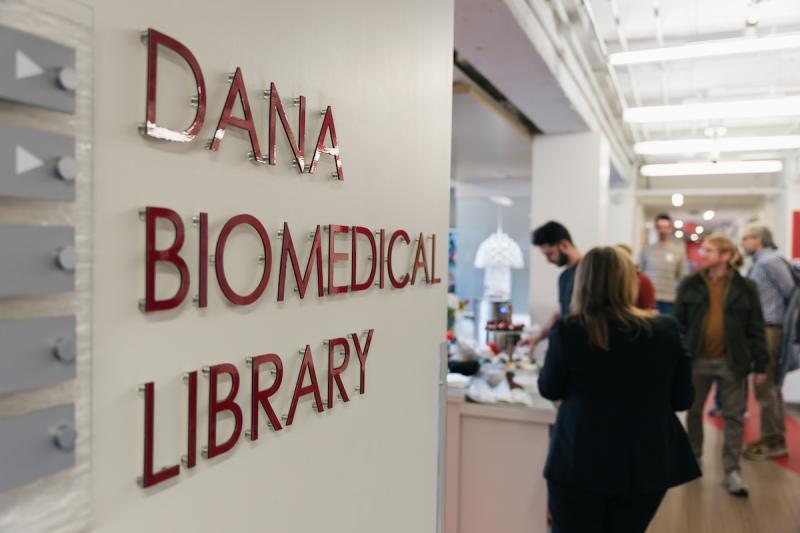Foreground shows wall text that reads 'DANA BIOMEDICAL LIBRARY' there are many people mingling in the kitchen in the background
