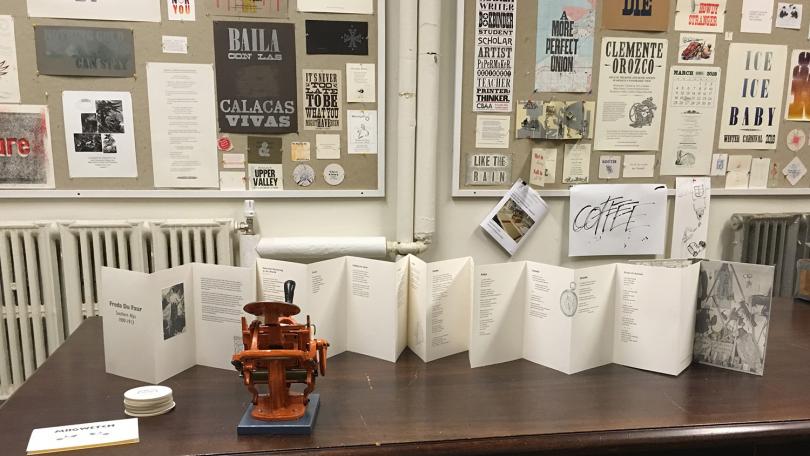 wall covered in printed posters, accordion book on table with tiny press