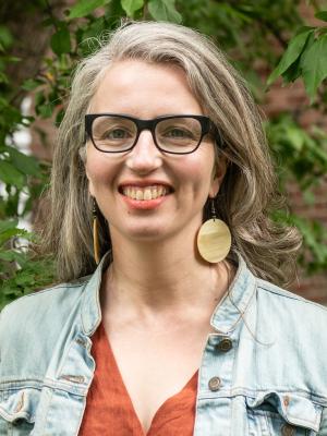 Picture of a woman wearing a light blue jacket, black rimmed glasses, and big round earrings