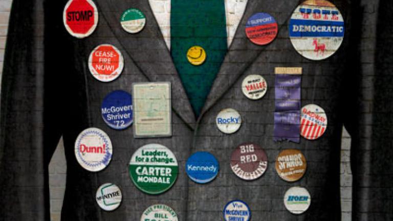 This exhibit poster has a suit jacket covered in an assortment of colorful political campaign pins and buttons representing multiple US elections and causes.