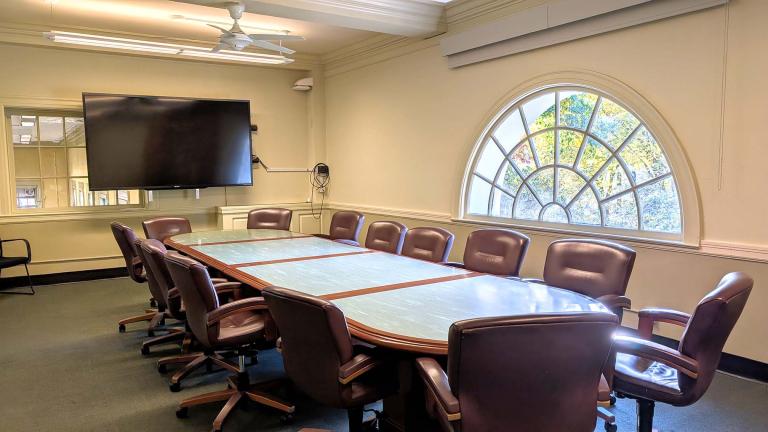 Conference table with chairs in a room with a mutlipaned window and a large flat screen display on the wall.